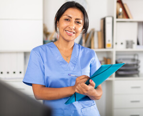Smiling Hispanic woman doctor in blue uniform standing in clinic, filling out clipboard with...