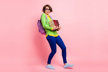 Full length photo of guilty young lady dressed yellow shirt glasses backpack walking holding book stack isolated pink color background