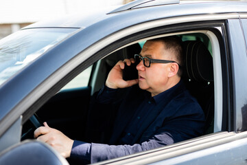 Cheerful man wearing eyeglasses sitting on the drivers seat and talking with his wife on the phone.