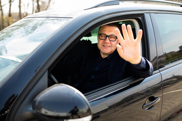 Mature man driving a car is happy and shows his satisfaction by smiling and raising his hand to greet