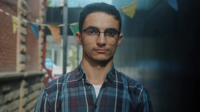 Slow motion portrait of attractive Middle-Eastern man wearing glasses standing outdoors in modern city and looking at camera with light smile