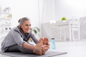 cheerful mature woman with grey hair stretching on yoga mat near sports bottle - Powered by Adobe