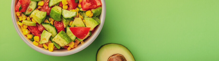 Tasty avocado salad with tomatoes, cucumbers in bowl on green background with copy space banner