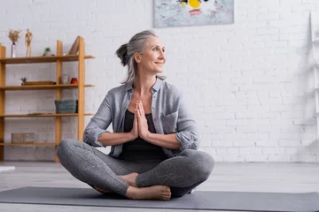  happy mature woman with grey hair sitting with praying hands in lotus pose on yoga mat © LIGHTFIELD STUDIOS
