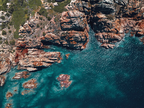 Aerial view of a breathtaking and wild coastline characterised by reddish cliffs and a turquoise transparent sea in Isola Rossa, Sardinia, Italy.