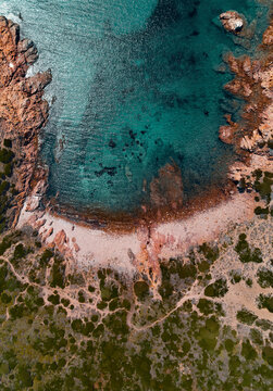 Aerial view of a beautiful and remote little beach surrounded by reddish cliffs and a turquoise transparent sea in Isola Rossa, Sardinia, Italy.