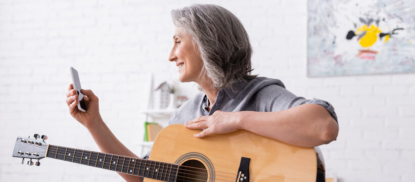 Smiling Mature Woman With Grey Hair Holding Smartphone While Learning To Play Acoustic Guitar, Banner