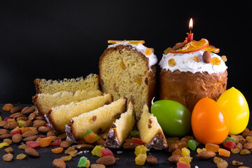 Traditional Easter cake on dark black background. Migdal, candied fruits, raisins, decor colorful eggs, candles are burning. Sectional view, slices, pieces. Orthodox treat. Copy space