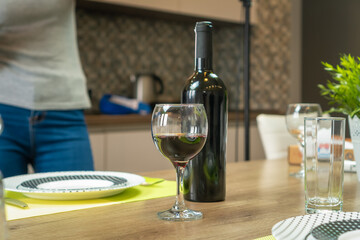 Glass of wine, a bottle, a plate, a glass stands on the kitchen table, in the background a girl in jeans and a T-shirt