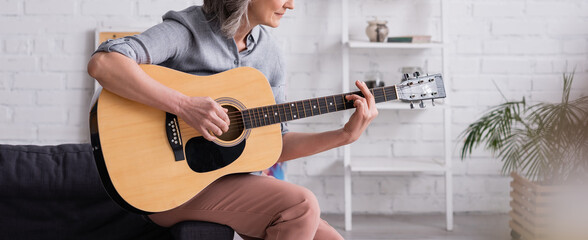 partial view of mature woman with grey hair playing acoustic guitar while sitting in living room,...