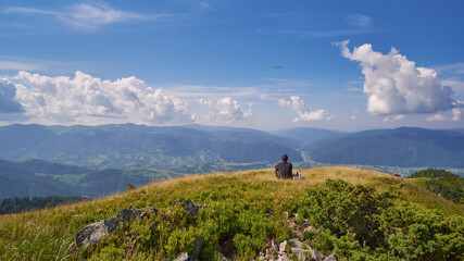 A lone tourist sits on the top of a mountain and surveys the valley. Sunny summer day, green grass everywhere. There are light clouds in the sky