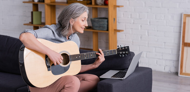 Happy Middle Aged Woman With Grey Hair Learning To Play Acoustic Guitar Near Laptop On Sofa, Banner