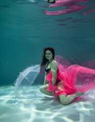 beautiful girl in a black swimsuit and with a pink pareo and with a pink umbrella underwater on a blue background 