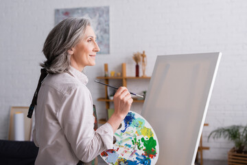 pleased middle aged woman holding paintbrush and palette with colorful paints near blank canvas