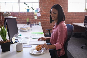 Mixed race businesswoman sitting in office in front of computer and having snack