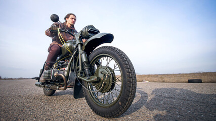 Close shoot of vintage motorcycle's wheel on the ride through old empty road at late autumn sunlight. Young old-fashioned motorcyclist in cool leather jackets looks hot in that journey.