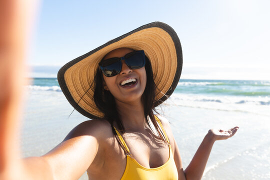 Smiling mixed race woman wearing sunhat and sunglasses taking selfie on beach