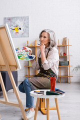 pensive mature artist holding paintbrush and palette while sitting near canvas