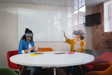 Diverse female colleagues sitting at work wearing virtual reality set and playing