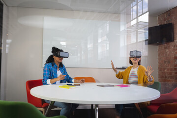 Diverse female colleagues sitting at work wearing virtual reality set and playing