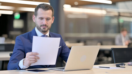 Middle Aged Businessman Reading Documents while using Laptop 