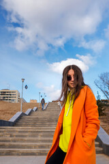 Obraz premium Active good-looking young woman in vivid outfit. Trendy female in sunglasses, orange coat, yellow hoodie and black jeans against the background of city stairs and blue sky.