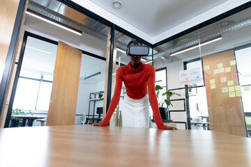 African american businesswoman leaning on desk and using vr headset in meeting room