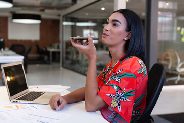 Portrait of mixed race businesswoman sitting in front of laptop having a call on smartphone