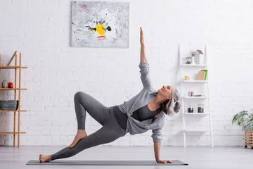 Fotobehang flexible middle aged woman in sportwear with raised hand practicing yoga on mat at home © LIGHTFIELD STUDIOS