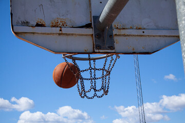 A basketball ball flies into a hoop with a metal net outdoors against a blue sky with white clouds...