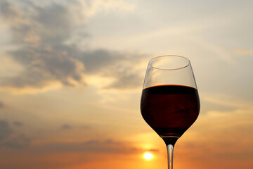 Glass with red wine on sunset background, evening sun and sky with clouds reflected in the glass. Concept of celebration, wine industry