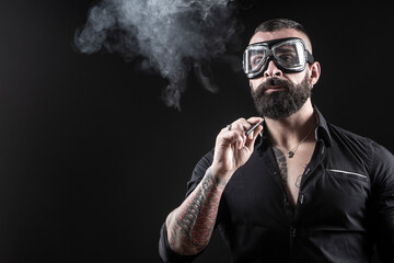 Uomo moro con la barba occhiali da motociclista,  fuma una sigaretta e produce una nuvola di fumo,...