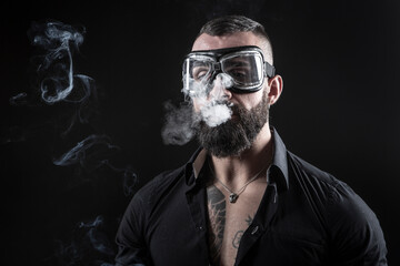 Uomo moro con la barba occhiali da motociclista,  fuma una sigaretta e produce una nuvola di fumo, isolato su sfondo nero