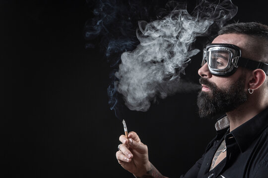 Uomo moro con la barba occhiali da motociclista,  fuma una sigaretta e produce una nuvola di fumo, isolato su sfondo nero