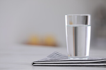 Glass of water and napkin on white table against blurred background. Space for text