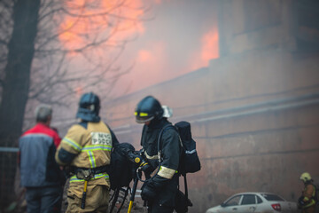 Fototapeta premium Firefighters put out large massive fire blaze, group of fire men in uniform during fire fighting operation in the city streets, firefighters with the fire engine truck fighting vehicle