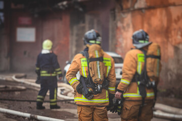 Fototapeta premium Firefighters put out large massive fire blaze, group of fire men in uniform during fire fighting operation in the city streets, firefighters with the fire engine truck fighting vehicle