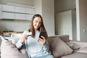 Convenience to do shopping without leaving home. Young charming woman holds smartphone and credit card sitting on the couch at home, making purchases, ordering food delivery, inputting card details