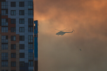 Fire fighting helicopter silhouette with bambi bucket for сarrying water to put out a massive building city fire, process of put out a large blaze bush fire wildifre, aerial firefighting with chopper