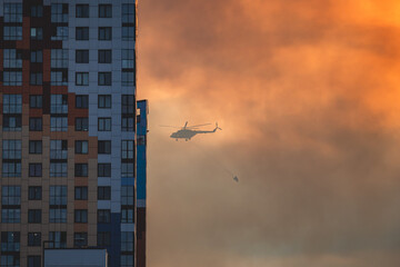 Fire fighting helicopter silhouette with bambi bucket for сarrying water to put out a massive building city fire, process of put out a large blaze bush fire wildifre, aerial firefighting with chopper