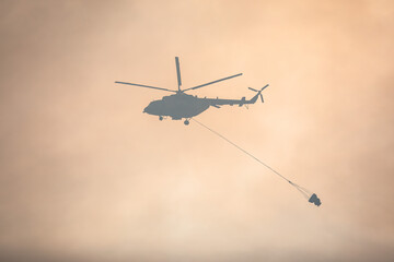 Fototapeta na wymiar Fire fighting helicopter silhouette with bambi bucket for сarrying water to put out a massive building city fire, process of put out a large blaze bush fire wildifre, aerial firefighting with chopper