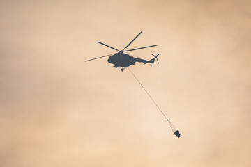 Fire fighting helicopter silhouette with bambi bucket for сarrying water to put out a massive building city fire, process of put out a large blaze bush fire wildifre, aerial firefighting with chopper