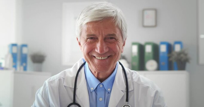 Senior doctor man sitting at desk in medical office looking at camera and smiling