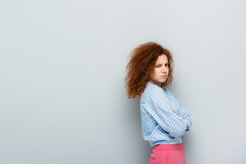 offended woman puffing cheeks while standing with crossed arms on grey background