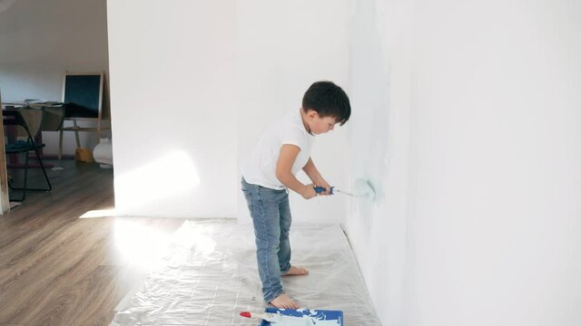 A boy paints a wall with a roller A Caucasian boy makes repairs at home A boy diligently paints over a white wall Side view