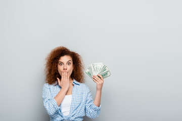 amazed woman covering mouth with hand while holding money on grey background