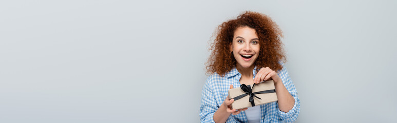 astonished woman looking at camera while opening present isolated on grey, banner