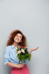 cheerful woman looking up and pointing with hand while holding flowers on grey background