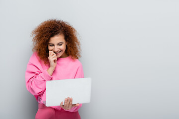 cheerful woman holding hand near face while looking at laptop on grey background