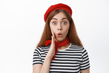 Close up of excited young girl in red beret and scarf, telling big secret or news, holding hand near mouth, talking to you, making announcement, standing over white background